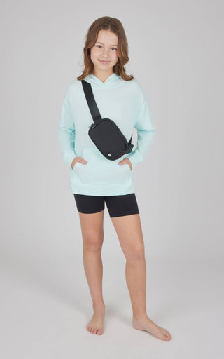 Young girl wearing a light blue hoodie and black shorts, holding a black crossbody bag on a plain background.