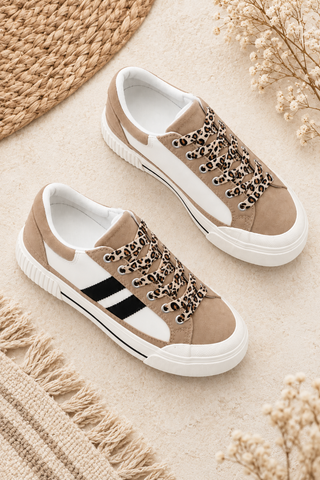 A pair of low-top canvas sneakers in beige and white, featuring leopard-print laces and a black side stripe, styled on a neutral beige surface with woven textures and dried flowers.
