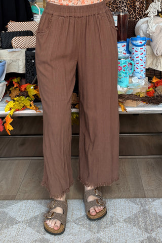 A person wearing loose, wide-leg brown pants with a frayed hem and double-strap sandals, standing indoors in a store or home setting with shelves and decor in the background.