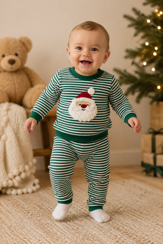 A baby wearing a green and white striped Christmas pajama set with a Santa face graphic on the chest, standing on a beige carpet in a cozy room with a teddy bear and a Christmas tree in the background.