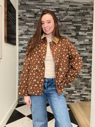 A woman wearing a brown button-up jacket with a white floral pattern, a cream ribbed top, and blue jeans, standing in front of a gray stone wall.