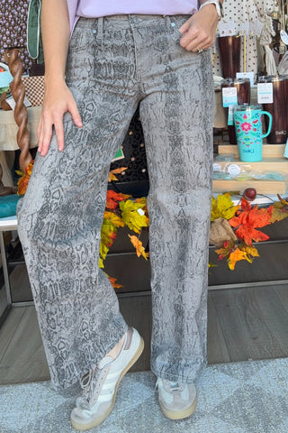 A person wearing gray snakeskin-print wide-leg jeans and white sneakers, standing in a retail store setting with shelves and fall decorations in the background.