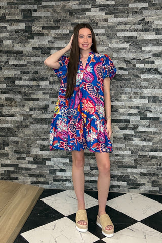 Woman wearing a colorful dress standing against a patterned wall.