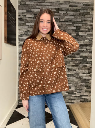 A woman modeling a brown long-sleeve button-up shirt with a beige and white floral pattern, paired with blue jeans, standing in front of a gray stone-tile wall.