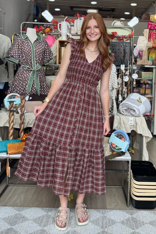 A woman with red hair stands in a retail store wearing a sleeveless, V-neck, tiered midi dress with a burgundy and brown plaid pattern. She is wearing beige sandals and standing in front of a busy background filled with other clothing and store fixtures.