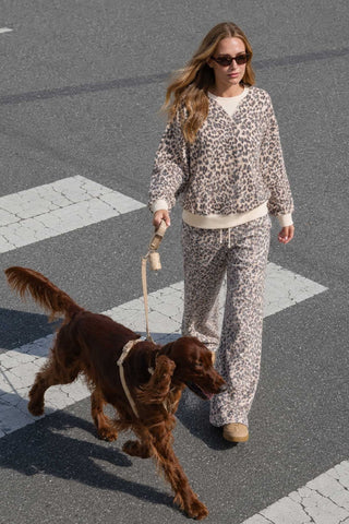 A woman walking a brown dog across a street crosswalk, wearing a matching beige and black leopard-print sweatshirt and pants set.