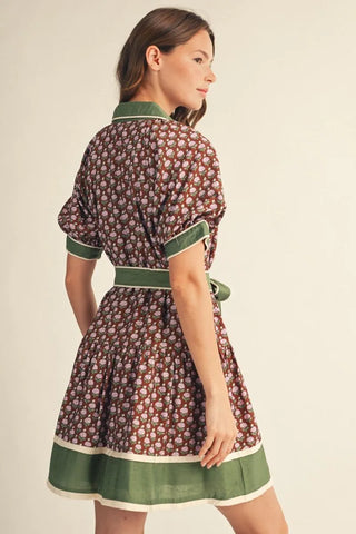 A woman modeling a short-sleeved mini dress from the back, featuring a brown and beige floral pattern with sage green trim, collar, cuffs, and a matching green waist tie, set against a plain beige background.