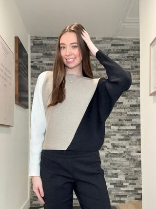 A woman stands in a hallway wearing a color-block knit sweater with a white long-sleeve base, a beige yoke, and black batwing sleeves, paired with black pants.