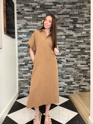 A woman modeling a tan short-sleeve midi shirt dress with a button-down collar, standing in front of a gray stone tile wall and a black-and-white checkered floor.