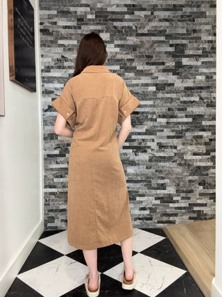 A woman viewed from above wearing a tan short-sleeve linen dress, standing on a black and white checkered floor against a gray stone tile wall.