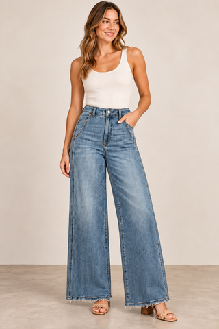 A woman modeling a pair of high-waisted, wide-leg blue denim jeans with a faded wash, styled with a white tank top and beige sandals against a neutral studio backdrop.