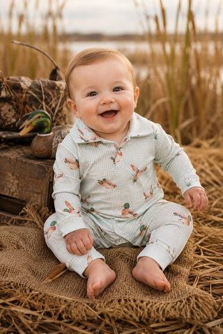 A smiling baby sitting outdoors on a burlap mat, wearing a light sage green long-sleeve romper with a small duck print. The background features a rustic wooden crate with a real mallard duck and tall golden reeds.