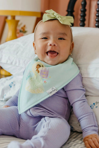 Baby wearing a purple outfit and green bib with a bow, sitting on a bed.