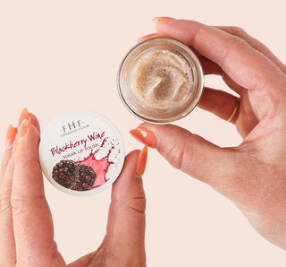 Jar of blackberry wine lip polish with a lid labeled 'Blackberry Wine' held by hands against a light background.