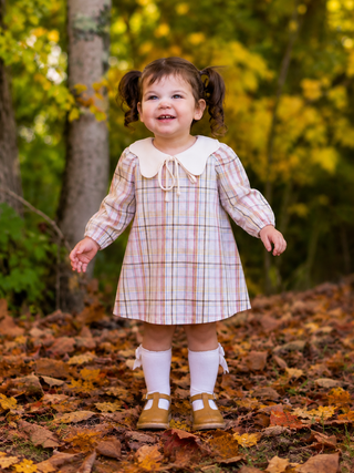 A young toddler girl with pigtails stands outdoors on a leaf-covered path wearing a cream-colored plaid dress with a large white Peter Pan collar, white socks, and tan Mary Jane shoes.