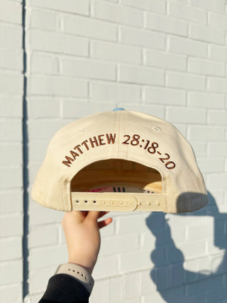 Beige cap with 'Matthew 28:18-20' text held against a white brick wall.