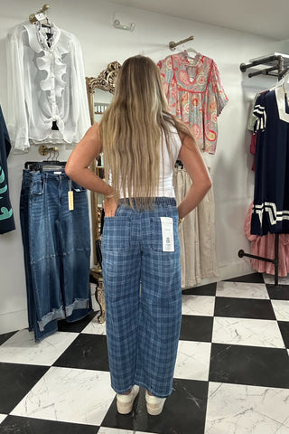 A woman stands with her back to the camera wearing blue plaid wide-leg pants, a white tank top, and white sneakers inside a clothing boutique. Clothing racks and a black-and-white checkered floor are visible in the background.