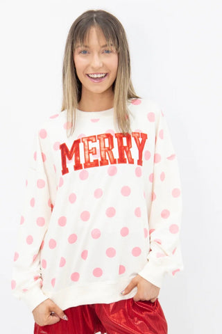 A woman modeling a white crewneck sweatshirt with pink polka dots and the word 'MERRY' in red varsity-style letters across the chest, paired with shiny red shorts, against a plain white background.