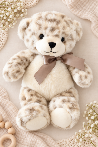 A plush teddy bear with cream and beige faux fur, brown spots, and a taupe ribbon bow, styled on a beige blanket with baby accessories.