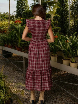 A woman stands with her back to the camera in a greenhouse or garden center, wearing a sleeveless red and burgundy plaid midi dress with ruffled cap sleeves and a smocked waist, paired with beige ankle boots.