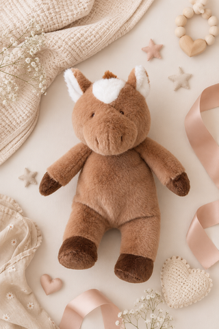 A plush brown horse toy with a white forehead patch and dark brown hooves, styled in a flat lay with a cream knit blanket, pink ribbons, wooden star toys, and baby accessories on a light beige background.