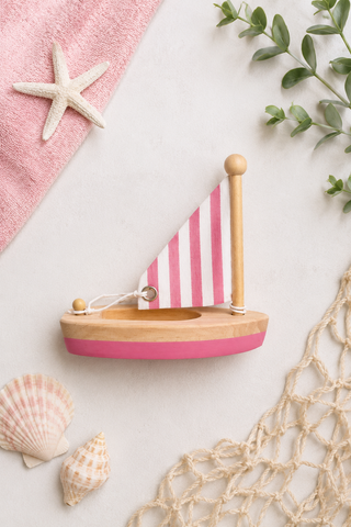 A wooden toy sailboat with a pink and white striped sail and a pink hull, styled on a light surface with a pink towel, starfish, shells, greenery, and netting.