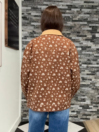 A person viewed from behind wearing a brown button-up shirt with a white floral pattern and a tan collar, paired with blue jeans, standing in front of a gray stone-tile wall.