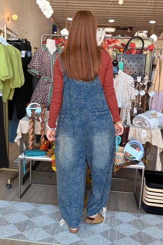 A woman viewed from behind wearing a pair of loose-fit blue denim overall jumpsuits over a rust-red long-sleeve shirt, standing inside a retail store.