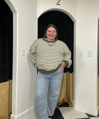 A woman wearing a green and white striped long-sleeve sweatshirt and light blue jeans, standing in a fitting room doorway with black curtains and white walls.