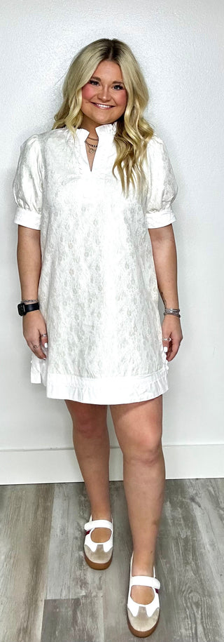 Woman wearing a white dress standing against a white wall.