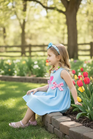 Young girl in a blue dress sitting on a brick ledge in a garden with tulips.