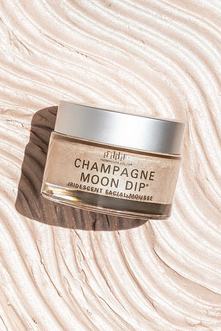 A jar of FARMHOUSE FRESH 'Champagne Moon Dip' iridescent facial mask resting on a textured, cream-colored background with wavy patterns.