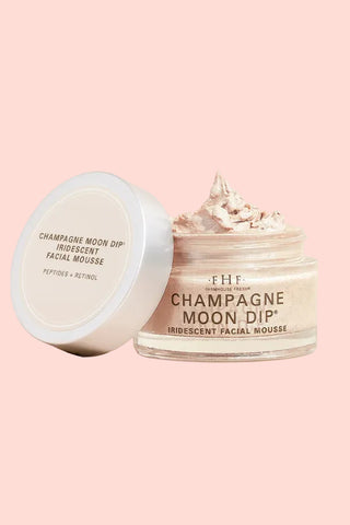 A clear cosmetic jar of The Champagne Moon Dip Iridescent Facial Mousse with its white lid leaning beside it on a warm beige background. The jar is open, showing whipped cream inside, with the lid displaying the product name in black text.