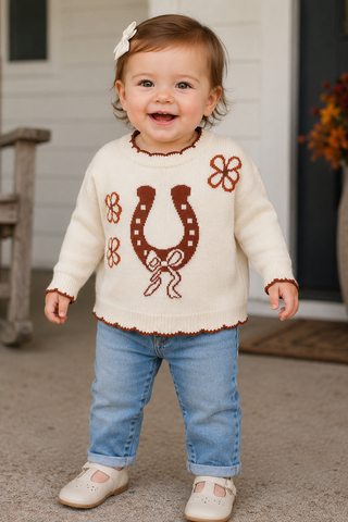 A toddler stands outdoors in front of a house door wearing a cream knit sweater with brown decorative motifs, blue jeans, and cream shoes. The sweater has a large brown horseshoe design on the front with small star details and matching brown floral accents.