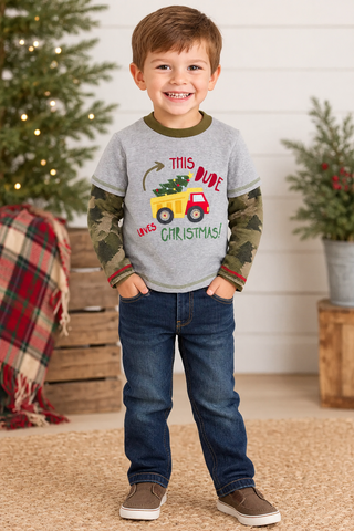A young boy stands smiling in a holiday-themed room, wearing a heather gray long-sleeve shirt with camouflage sleeves and a Christmas graphic that reads 'This Dude Likes Christmas!' with a yellow truck carrying a green Christmas tree. He is also wearing blue jeans and brown shoes.