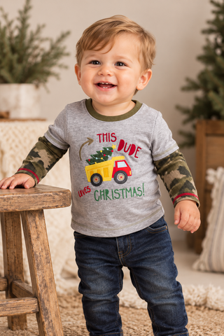 A toddler standing indoors wearing a heather gray long-sleeve shirt with camouflage sleeves and a Christmas truck graphic that reads 'THIS DUDE LOVES CHRISTMAS!'. The child is also wearing dark blue jeans and smiling while leaning on a wooden stool.