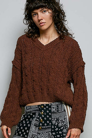 A model wearing a brown, textured, long-sleeve cropped sweater and black-and-white patterned pants against a plain gray background.
