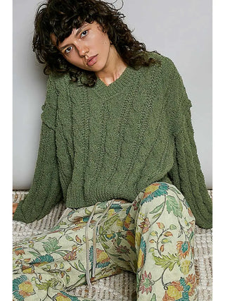 A woman wearing an oversized textured olive green knit sweater and floral print pants, sitting on a woven bench against a plain light gray wall.