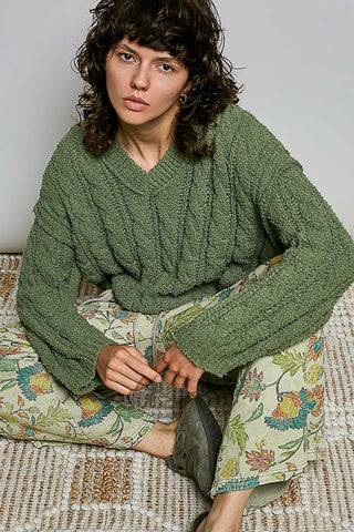 A woman wearing a chunky olive green cable-knit cropped sweater and light green floral print pants, photographed against a plain light gray background.