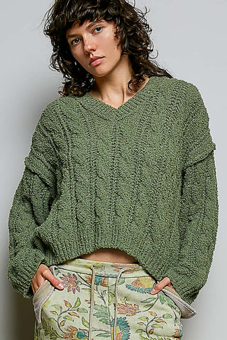 A woman wearing a chunky olive green cable-knit cropped sweater and light green floral print pants, photographed against a plain light gray background.