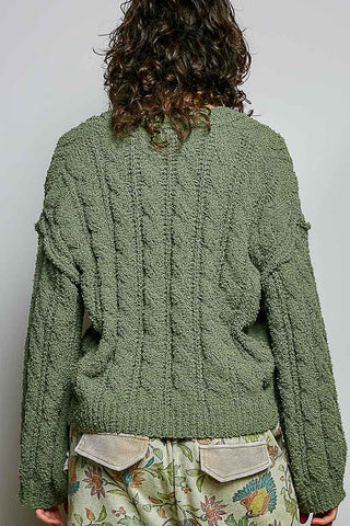 A person shown from the back wearing an olive green chunky cable-knit cardigan and a floral print skirt with beige patch pockets, against a plain light gray background.