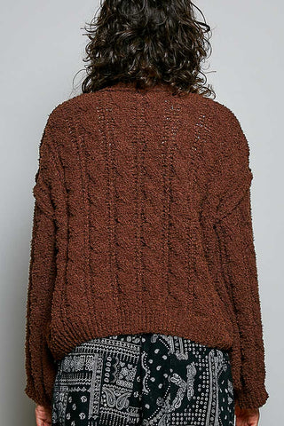 A person viewed from the back wearing a chunky, textured brown cable-knit sweater and black-and-white patterned pants against a plain light gray background.