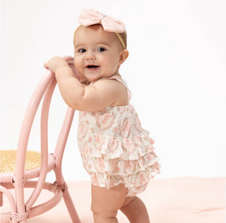 Baby in a floral romper with a pink bow sitting on a pink chair against a white background