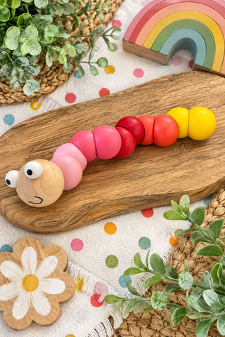 A wooden caterpillar toy with colorful beads in pink, red, orange, and yellow, displayed on a wooden board with nursery-themed props.