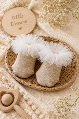 A pair of baby booties made of natural burlap with white tulle ruffles, displayed in a woven oval basket on a cream knit blanket with baby props including a wooden heart rattle and a wooden sign that reads 'little blessing'.