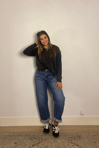 Woman wearing a black sweater and blue jeans standing against a plain wall.