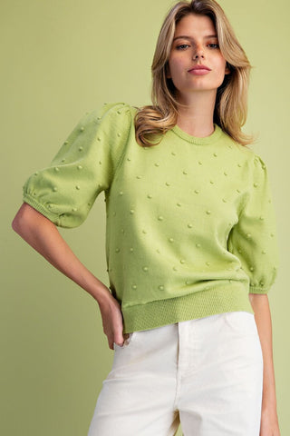 A woman wearing a light lime green knit sweater with textured 3D puff sleeves and white pants, posing against a solid light green background.