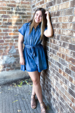 A woman standing with one hand against a brick wall, wearing a blue denim romper with button-down detailing and a flowy fit.