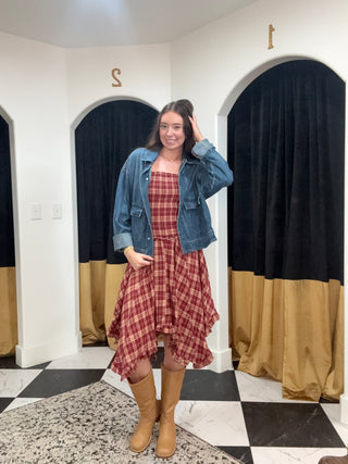 A woman stands in a fitting room wearing a red and white plaid strapless dress with a blue denim jacket and tan knee-high boots. The background features white walls, black and gold curtains, and a black-and-white checkered floor.