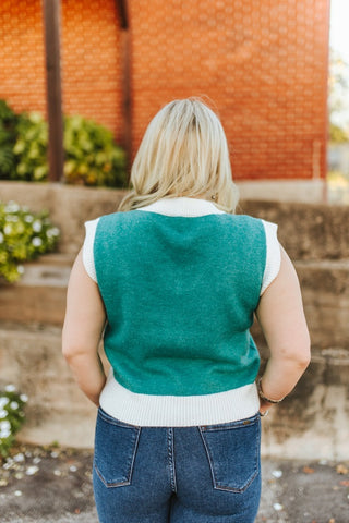 Person wearing a green vest over a white shirt with blue jeans, standing outdoors.